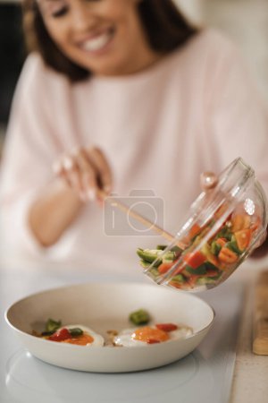Téléchargez les photos : Tourné d'une jeune femme préparant des œufs frits avec des légumes pour le petit déjeuner dans sa cuisine. Concentration sélective. - en image libre de droit