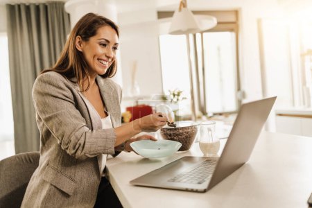 Téléchargez les photos : Multi-tâches jeune femme d'affaires en utilisant son ordinateur portable et prendre un petit déjeuner tout en se préparant à aller au travail. - en image libre de droit