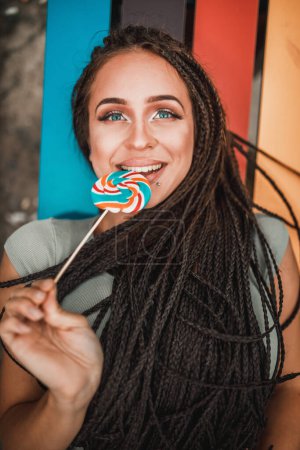 Photo for Young woman with long afro braids having fun and licking the lollipop on the colorful bench in the park. - Royalty Free Image