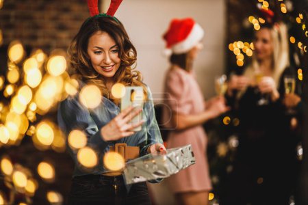 Téléchargez les photos : Souriant jeune femme tenant présent et prenant un selfie avec smartphone lors de la fête du Nouvel An à la maison. - en image libre de droit