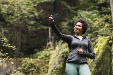 Photo for Mature African American woman making a video call while standing near the waterfall and enjoying the view during her hike in the mountains. - Royalty Free Image