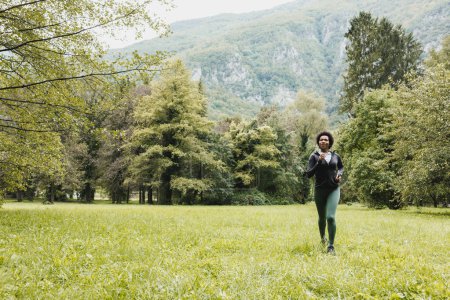 Foto de Hermosa sonriente mujer afroamericana madura está corriendo a través de hierba verde cerca del paisaje de montaña. - Imagen libre de derechos