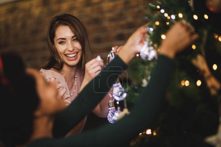 Foto de Hermosas amigas multiétnicas disfrutando de las vacaciones, decorando el árbol de Navidad, arreglando las luces de Navidad y divirtiéndose. - Imagen libre de derechos