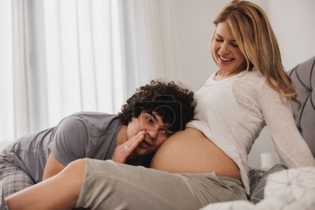 Foto de Feliz pareja embarazada divirtiéndose y comunicándose con el bebé embarazada en el estómago de la madre embarazada en casa. - Imagen libre de derechos