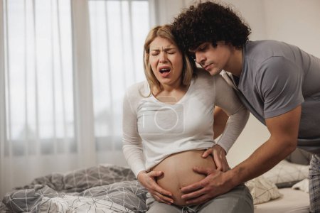 Téléchargez les photos : Homme inquiet aidant sa femme enceinte dans une chambre à coucher qui a des contractions prénatales. - en image libre de droit