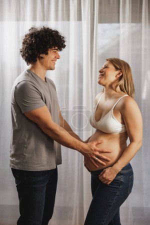 Téléchargez les photos : Joyeux couple enceinte amoureux parlant tout en se tenant près de la fenêtre à la maison. - en image libre de droit