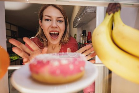 Photo for View looking out from inside of refrigerator as woman opens door and excited reaches for unhealthy donut. Selective focus. - Royalty Free Image