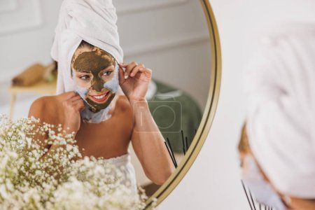 Téléchargez les photos : Belle jeune femme devant le miroir appliquant un masque facial au salon de beauté. - en image libre de droit