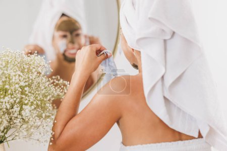 Téléchargez les photos : Belle jeune femme en face du miroir enlever masque facial dans la salle de bain à la maison. - en image libre de droit