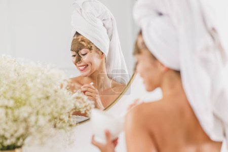 Foto de Hermosa joven frente al espejo aplicando mascarilla facial en el baño en casa. - Imagen libre de derechos