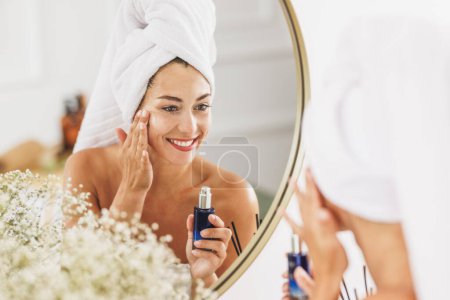 Téléchargez les photos : Belle jeune femme devant le miroir appliquant de l'hydratant sur son visage au salon de beauté. - en image libre de droit