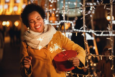 Photo for Middle age black woman holding sparklers and Christmas present box at festive winter night in the city. - Royalty Free Image