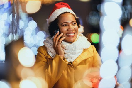 Photo for Middle age African woman talking on smartphone on the Christmas market at festive night in the city. - Royalty Free Image