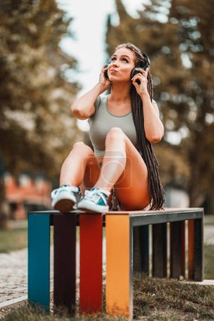 Foto de Linda mujer joven con el pelo largo trenzado afro escuchar música de un auricular mientras está sentado en el banco de colores en el parque. - Imagen libre de derechos