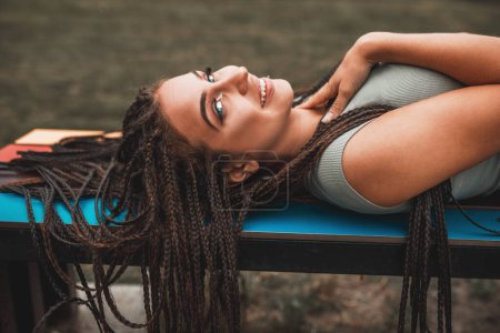 Téléchargez les photos : Mignon jeune femme avec de longues tresses afro profiter tout en étant couché sur le banc coloré dans le parc et coûteux regarder la caméra. - en image libre de droit