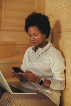 Téléchargez les photos : Femme noire assise dans les escaliers et utilisant smartphone et ordinateur portable tout en passant du temps de vacances dans une maison en bois confortable dans une forêt. - en image libre de droit