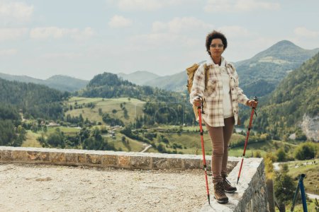 Photo for Mature black woman with backpack and trekking poles standing in nature and enjoying in fresh air while hiking on mountain. - Royalty Free Image