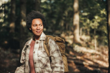 Foto de Mujer negra madura con mochila disfrutando de la vista del bosque mientras camina en la naturaleza. - Imagen libre de derechos