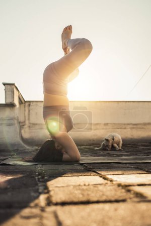 Téléchargez les photos : Relaxed woman practicing yoga headstand on a rooftop terrace at sunset. - en image libre de droit