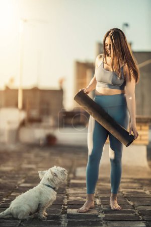 Téléchargez les photos : Femme détendue prépare tapis d'exercice pour pratiquer le yoga sur une terrasse sur le toit soutenant par son chien de compagnie au coucher du soleil. - en image libre de droit