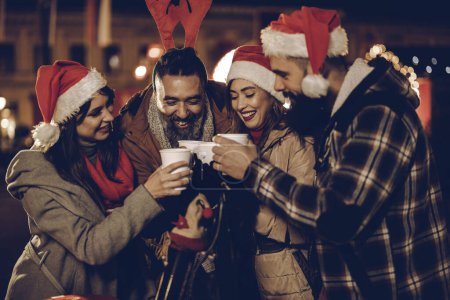 Téléchargez les photos : Tournage d'un groupe de jeunes amis joyeux s'amusant ensemble et dégustant du vin chaud sur le marché de Noël lors d'une soirée. - en image libre de droit