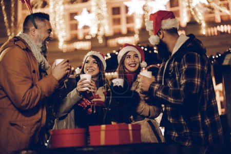Foto de Foto de un grupo de alegres jóvenes amigos divirtiéndose juntos y disfrutando del vino caliente en el mercado de Navidad en una fiesta nocturna. - Imagen libre de derechos