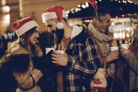 Téléchargez les photos : Tournage d'un beau jeune couple dégustant du vin chaud sur le marché de Noël lors d'une soirée. - en image libre de droit
