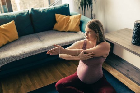 Foto de Mujer embarazada joven haciendo yoga en casa. Ella está haciendo ejercicios de estiramiento en su sala de estar. - Imagen libre de derechos