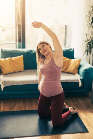 Téléchargez les photos : Jeune femme enceinte faisant de l'exercice à la maison le matin. Elle fait de l'exercice d'étirement. - en image libre de droit