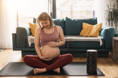 Photo for A pregnant woman in sports clothing is relaxing and exercising to promote wellbeing in her living room. - Royalty Free Image