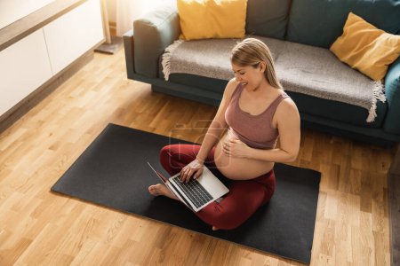 Photo for Young pregnant woman using laptop while exercising at home in the morning. She relaxing on exercise mat. - Royalty Free Image