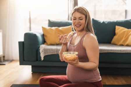 Foto de Feliz mujer embarazada comiendo ensalada de frutas frescas mientras hace ejercicio en casa. Ella se relaja en la alfombra de ejercicio en su sala de estar por la mañana. - Imagen libre de derechos