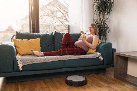 Photo for Young pregnant woman relaxingat on sofa at home in the morning while robot vacuum cleaner is working and cleaning living room. - Royalty Free Image