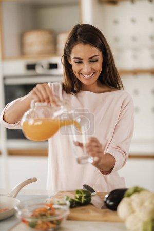 Téléchargez les photos : Tourné d'une jeune femme souriante versant et buvant du jus d'orange frais dans sa cuisine. - en image libre de droit