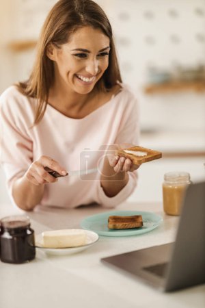 Photo for Shot of a young smiling woman making video call on a laptop while preparing breakfast for herself in her kitchen. - Royalty Free Image