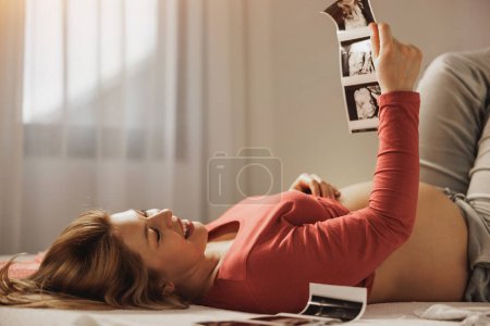 Foto de Mujer embarazada joven relajada mirando el ultrasonido de su bebé mientras se relaja en una cama en el dormitorio. - Imagen libre de derechos