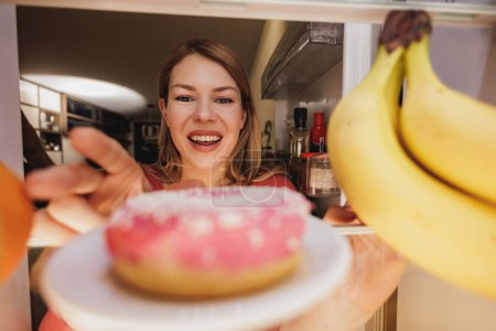 Foto de Vista desde el interior del refrigerador mientras la mujer abre la puerta y se emociona por la rosquilla poco saludable. Enfoque selectivo. - Imagen libre de derechos