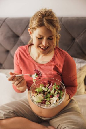 Téléchargez les photos : Heureuse femme qui attend de manger de la salade de légumes frais tout en étant assise sur le lit dans sa chambre. - en image libre de droit