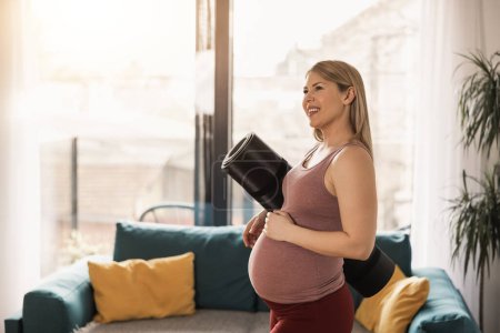 Téléchargez les photos : Femme enceinte tenant tapis d'exercice est prêt pour la pratique du yoga à la maison le matin. - en image libre de droit