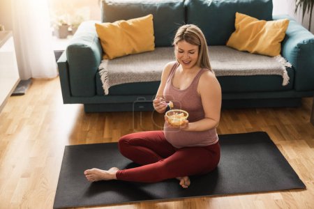 Téléchargez les photos : Joyeux jeune femme enceinte manger de la salade de fruits frais tout en faisant de l'exercice à la maison. Elle se relaxe sur un tapis d'exercice dans son salon le matin. - en image libre de droit