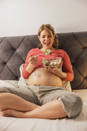 Téléchargez les photos : Heureuse femme enceinte manger de la salade de légumes frais tout en étant assis sur le lit dans sa chambre. - en image libre de droit