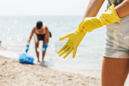 Foto de Close-up of a unrecognizable female volunteer putting protective rubber yellow gloves while cleaning sea beach area from the trash. - Imagen libre de derechos
