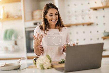 Photo for Shot of a young smiling woman using laptop to make a video chat with someone while preparing a healthy meal at home. - Royalty Free Image