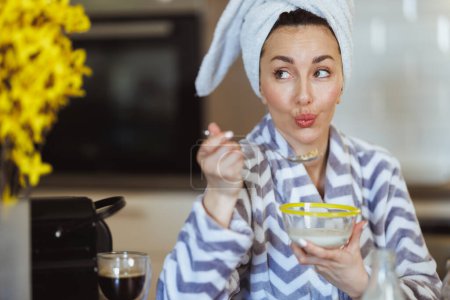 Téléchargez les photos : Une jolie jeune femme en peignoir mangeant des céréales pour le petit déjeuner à la maison. - en image libre de droit