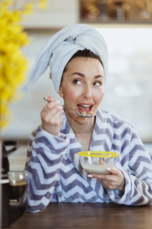 Foto de Mujer joven en albornoz comiendo cereales para el desayuno y teniendo ritual de placer matutino en casa. - Imagen libre de derechos