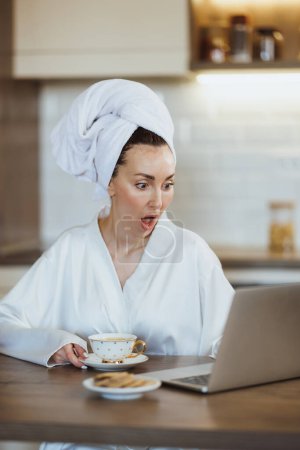 Photo for Young surprised woman in bathrobe surfing the at on her laptop while drinking a morning coffee at home. - Royalty Free Image