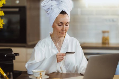 Photo for Young woman in bathrobe paying bills with credit card while working on her laptop in the her kitchen. - Royalty Free Image