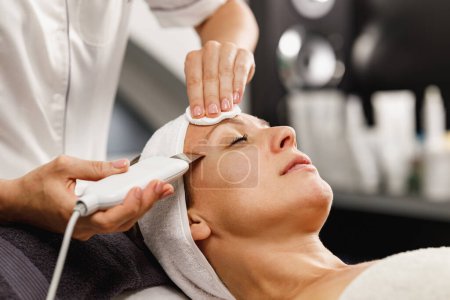Téléchargez les photos : Tournage d'une belle femme mature sur un traitement facial ultrasonique au salon de beauté. - en image libre de droit