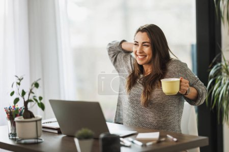 Téléchargez les photos : Jeune femme pigiste profiter d'une tasse de café tout en travaillant à partir de son bureau à domicile. - en image libre de droit