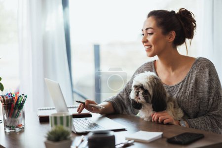 Foto de Atractiva joven freelancer trabajando en el portátil desde su casa y teniendo a su perro mascota en su regazo para hacerle compañía. - Imagen libre de derechos